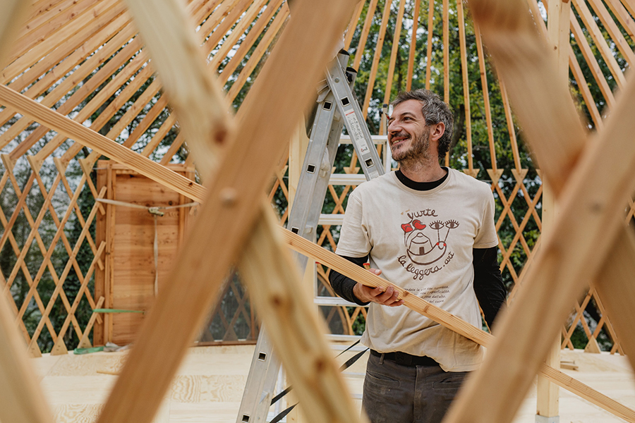 Uomo sorridente mentre costruisce una yurta in legno, con una scala sullo sfondo e la struttura parzialmente completata.
