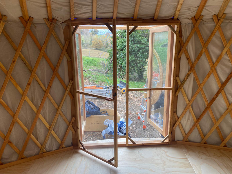 Interno di una yurta con porta in legno aperta, mostrando attrezzi da lavoro e paesaggio naturale esterno.