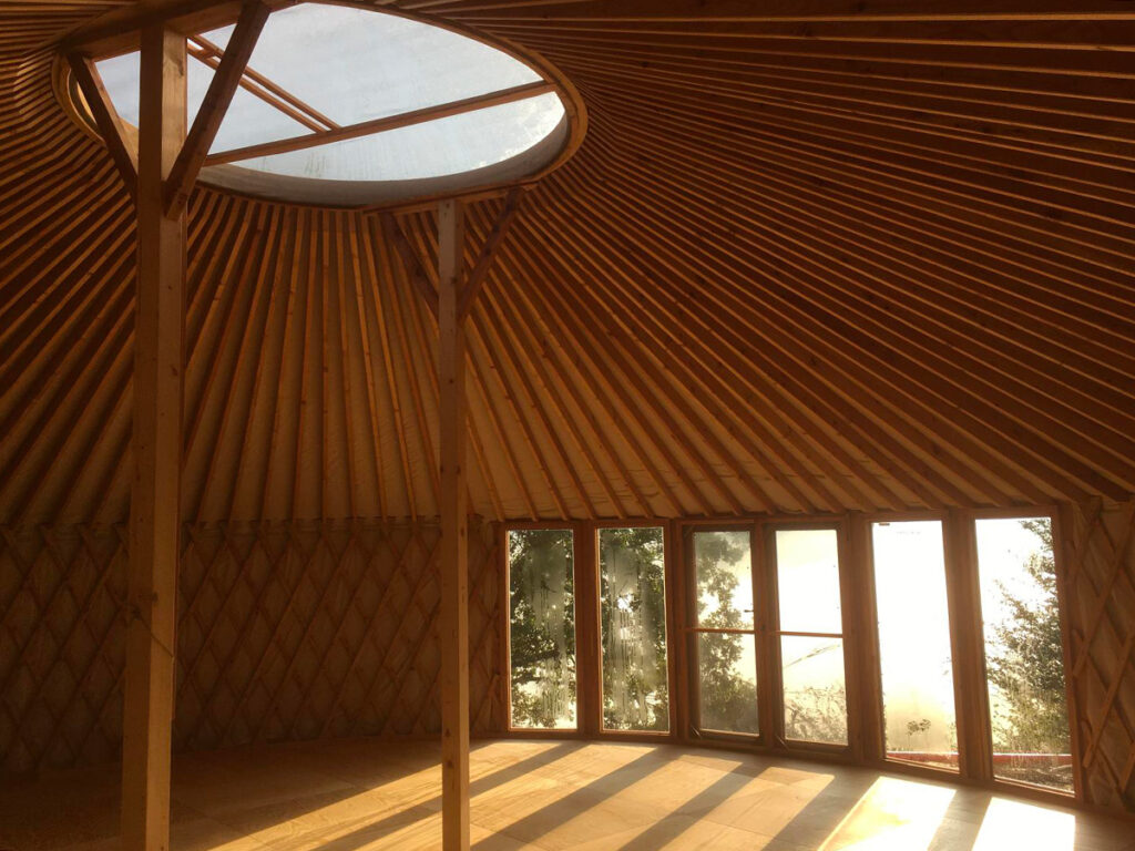 Interno di una yurta in legno con ampie finestre e cupola trasparente, illuminata dalla luce naturale del tramonto